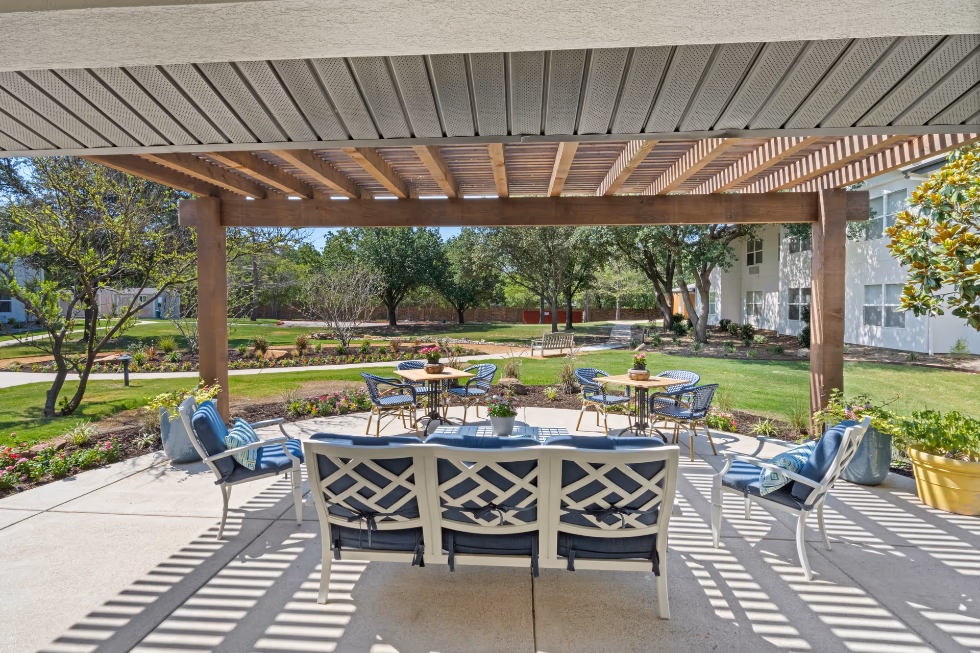 Outdoor patio area with a wooden pergola providing partial shade. The patio has cushioned chairs and a loveseat arranged around a small table, with additional tables and chairs in the background. Surrounding the patio is a well-maintained garden with green grass, trees, and flowering plants. A building with white walls and windows is visible on the right side.