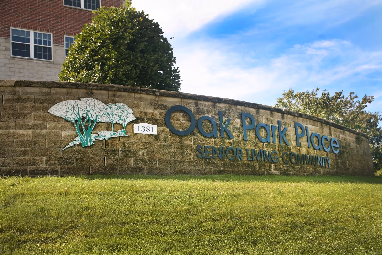 Stone entrance sign reading 'Oak Park Place Senior Living Community' with the address '1381' on a grassy slope in front of a building and trees.