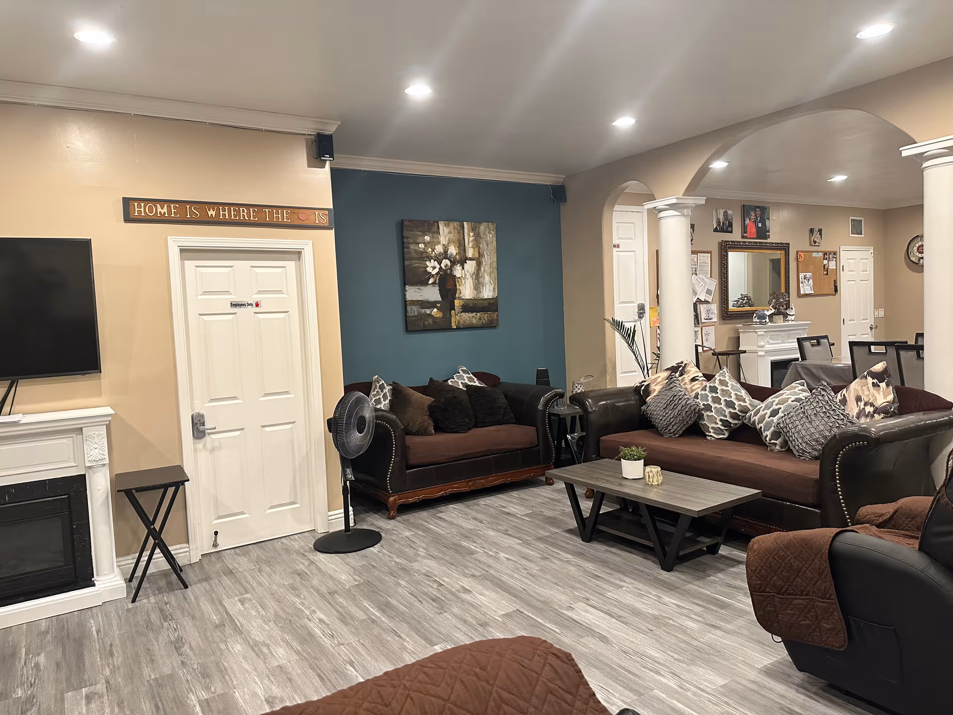 A cozy living room in a senior residence featuring two brown leather sofas with multiple patterned cushions, a wooden coffee table with small decorative items, a standing fan, a wall-mounted TV, and a white door labeled 'Employees Only'. The room has light-colored walls with one accent wall painted teal, a painting of flowers, and white columns separating the living area from a dining space with a table and chairs. The floor is covered with light gray wood-like flooring.