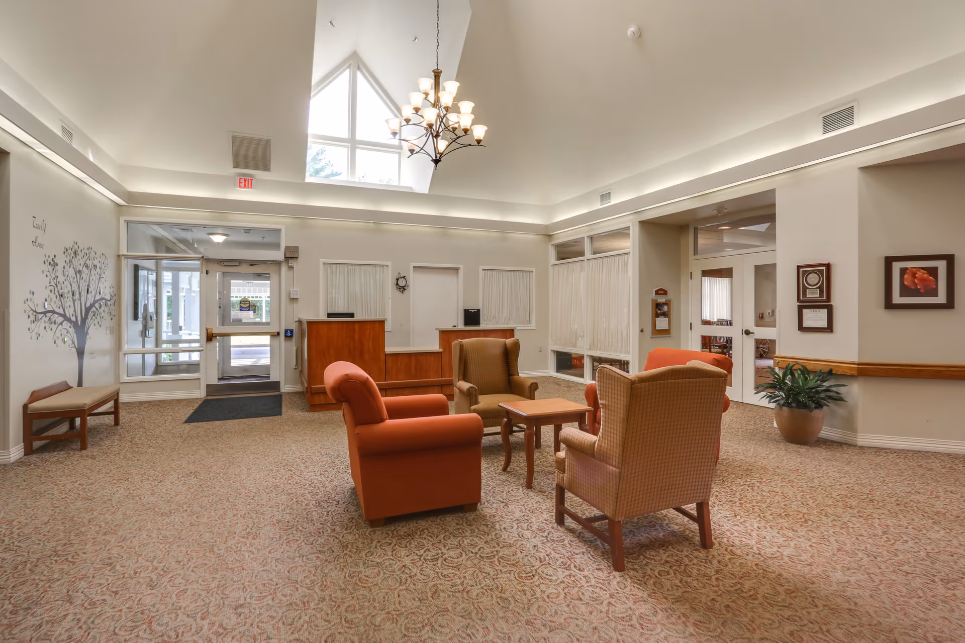 A spacious and well-lit common area in a senior living facility with a high vaulted ceiling and a chandelier. The room features a seating arrangement with four armchairs around a small wooden table on a patterned carpet. There is a reception desk against the far wall, large windows, a glass door entrance, a bench, a potted plant, and framed artwork on the walls.