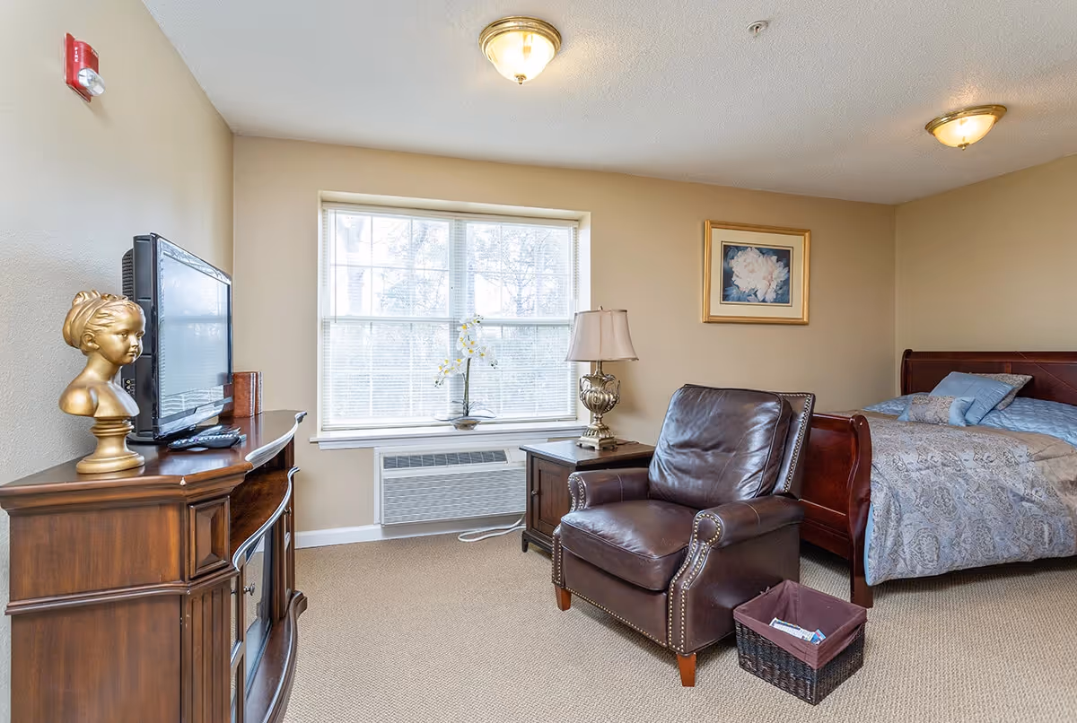 A cozy senior living room and bedroom area with a large window letting in natural light. The room features a dark leather armchair, a wooden side table with a decorative lamp, a wooden TV stand with a flat-screen TV and a golden bust sculpture. The bed has a wooden frame and is made with patterned bedding and pillows. A framed floral painting hangs on the beige wall.