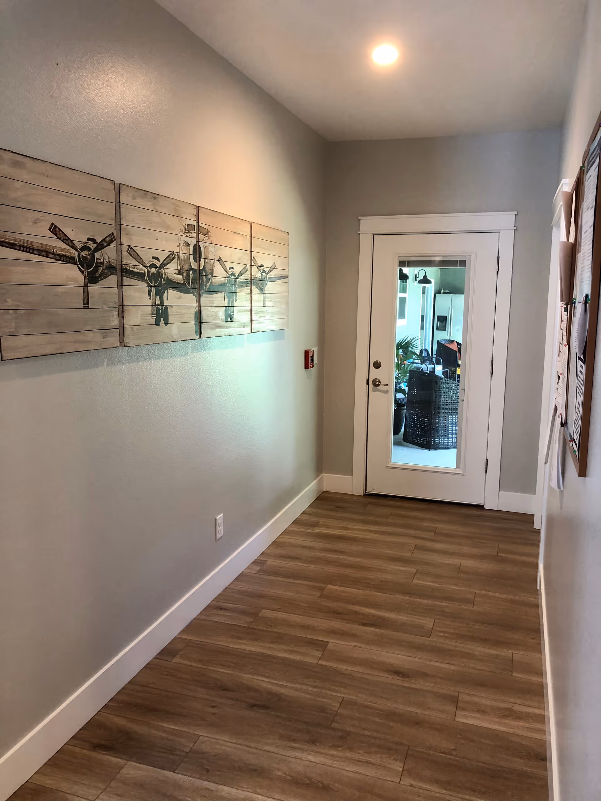A hallway with light gray walls and wood-look flooring leading to a white door with a glass panel. On the left wall, there is a four-panel artwork depicting an airplane. On the right wall, there is a bulletin board with papers pinned to it. The hallway is lit by a ceiling light.