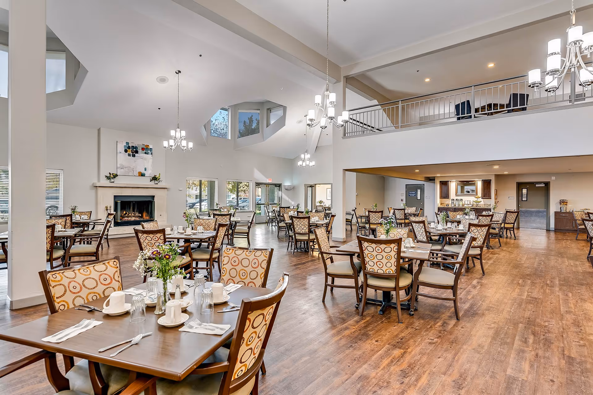 Large, airy dining room with multiple set tables and chairs, chandeliers, a fireplace, and a second-floor balcony.