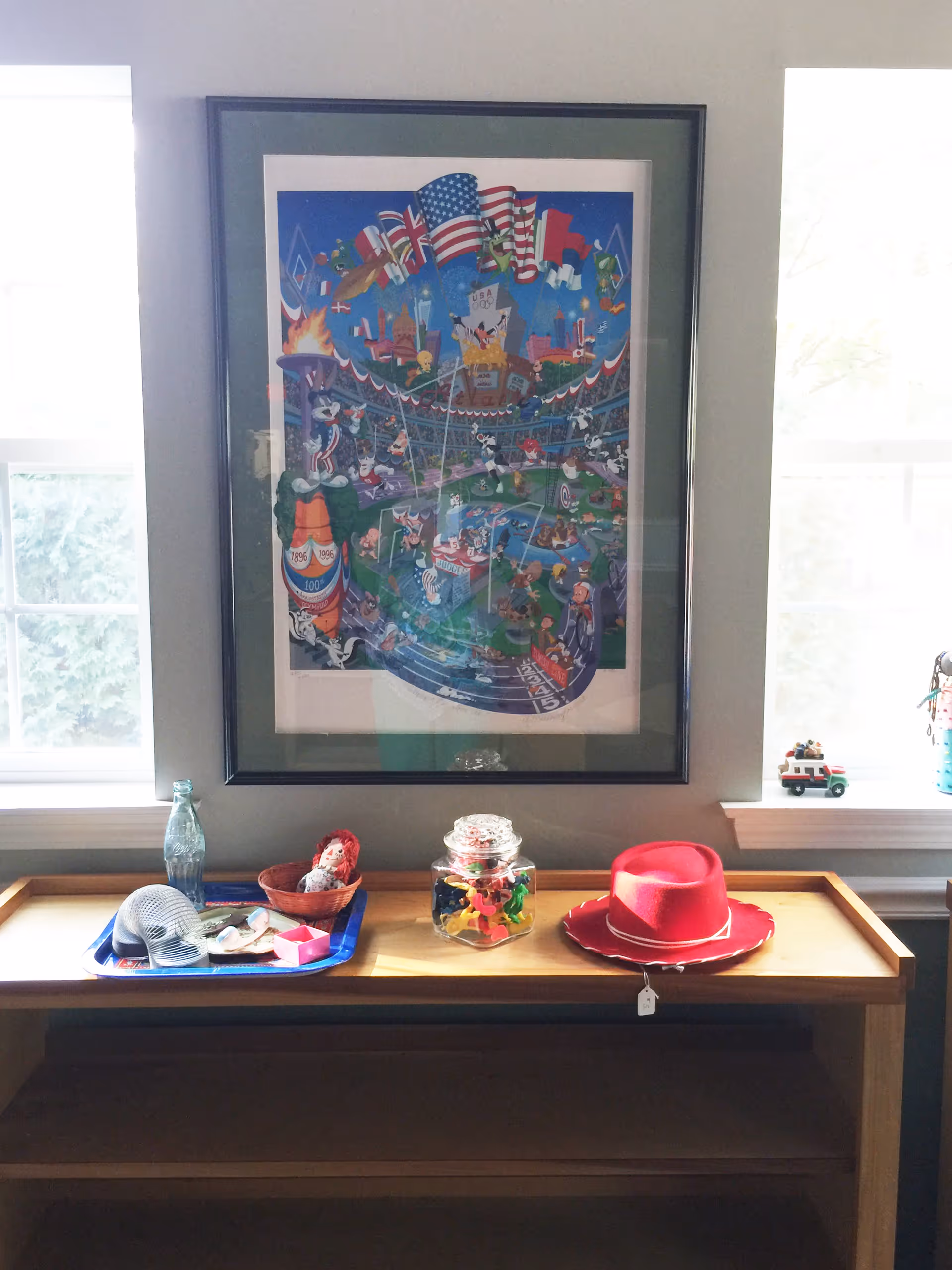 A wooden table with various items on it including a blue tray holding a metal mesh hat, a small doll in a basket, a glass bottle, a jar filled with colorful plastic toys, and a red cowboy hat with a price tag. Above the table is a framed colorful cartoon poster featuring various animated characters and flags. The table is positioned between two windows with white frames.