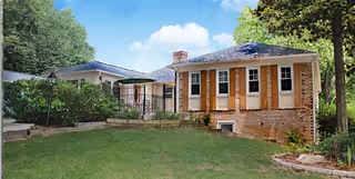 Exterior view of a single-story brick building with multiple windows and a shingled roof, surrounded by green grass, trees, and shrubs under a blue sky with some clouds.
