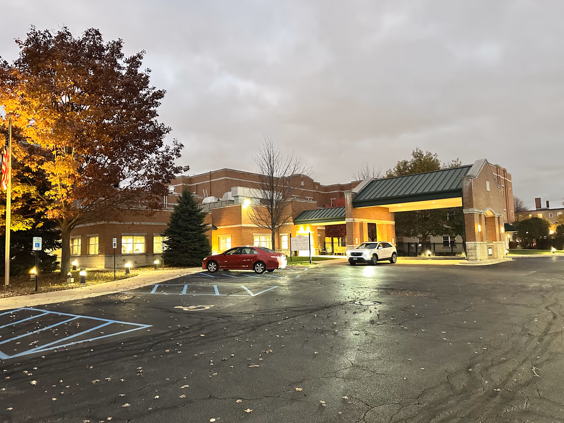 Exterior front entrance of a brick senior living building at dusk with a covered drive-through, parked cars, and illuminated windows.