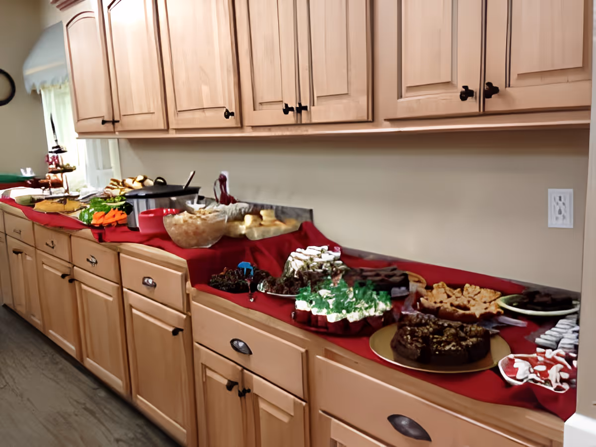 A long kitchen counter with wooden cabinets above and below, covered with a red tablecloth and an assortment of food items including vegetables, dips, desserts, and baked goods arranged for a buffet-style serving.