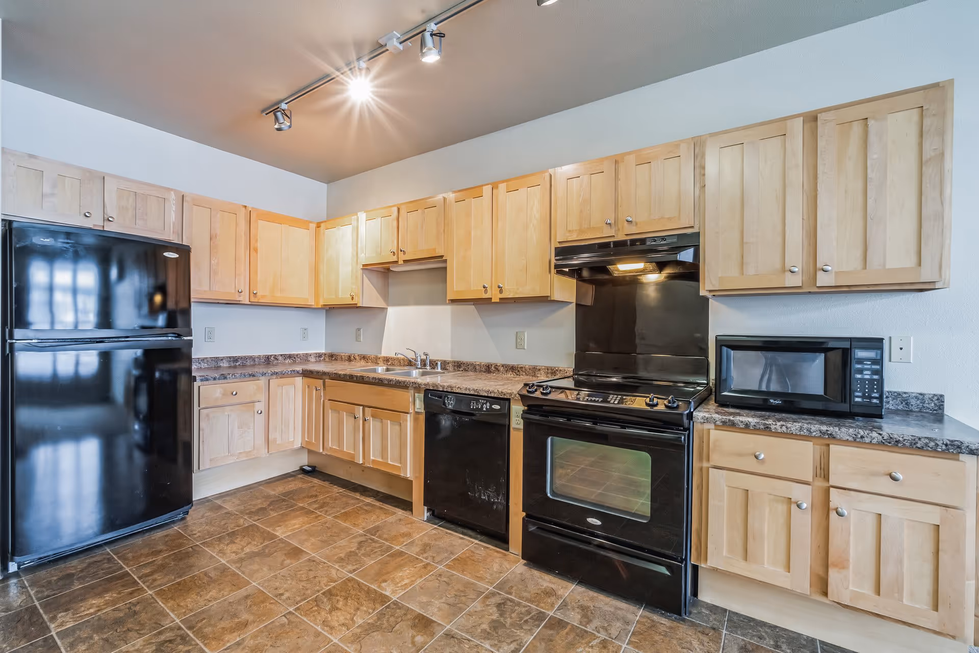 Modern kitchen with light wooden cabinets, black refrigerator, oven, dishwasher and microwave, a double sink and tiled floor.