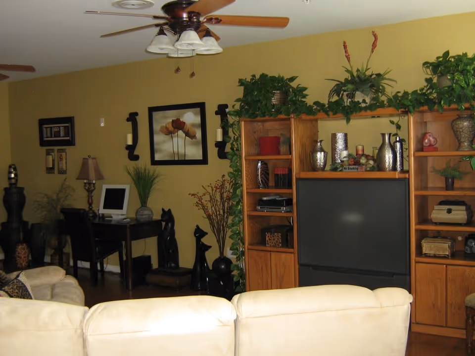 A cozy living room with beige sofas facing a large wooden entertainment center that holds a television and various decorative items including vases and plants. The walls are painted yellow, and there is a ceiling fan with lights. To the left, there is a small desk with a computer and chair, along with framed artwork and additional decorative plants and sculptures.