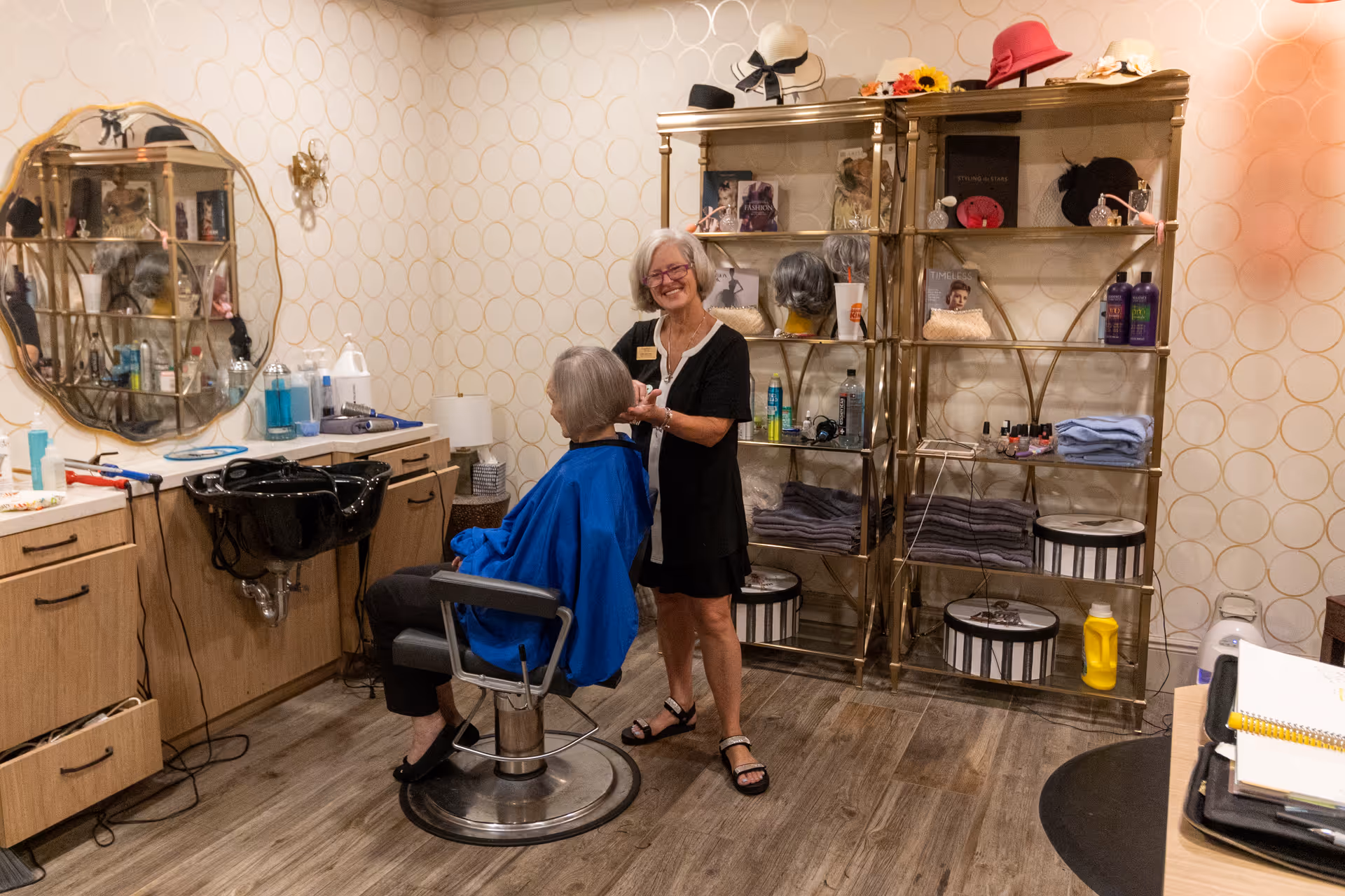 A hair salon room with a hairstylist standing and smiling while working on an elderly woman's hair. The woman is seated in a salon chair wearing a blue cape. The room has wooden flooring, a round mirror on the wall, shelves with hair products, hats, and towels, and a black hair washing sink attached to a wooden cabinet.