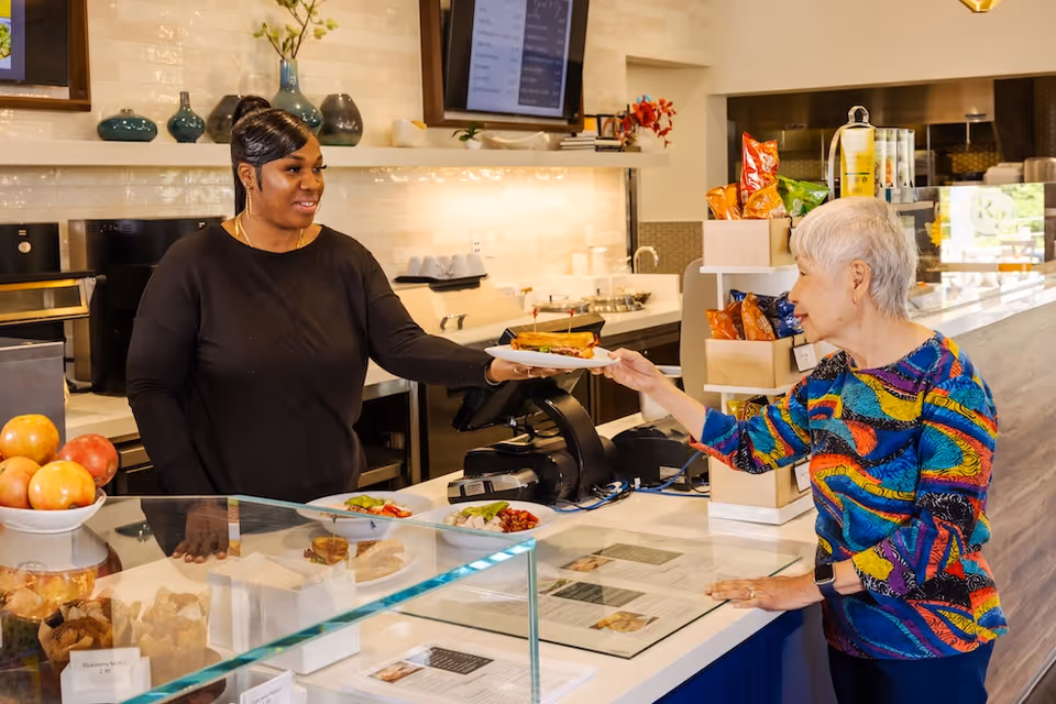 A woman behind a counter hands a plate with a sandwich to an elderly woman wearing a colorful shirt in a bright cafe or dining area. The counter displays various food items and snacks, with a bowl of apples and a cash register visible.