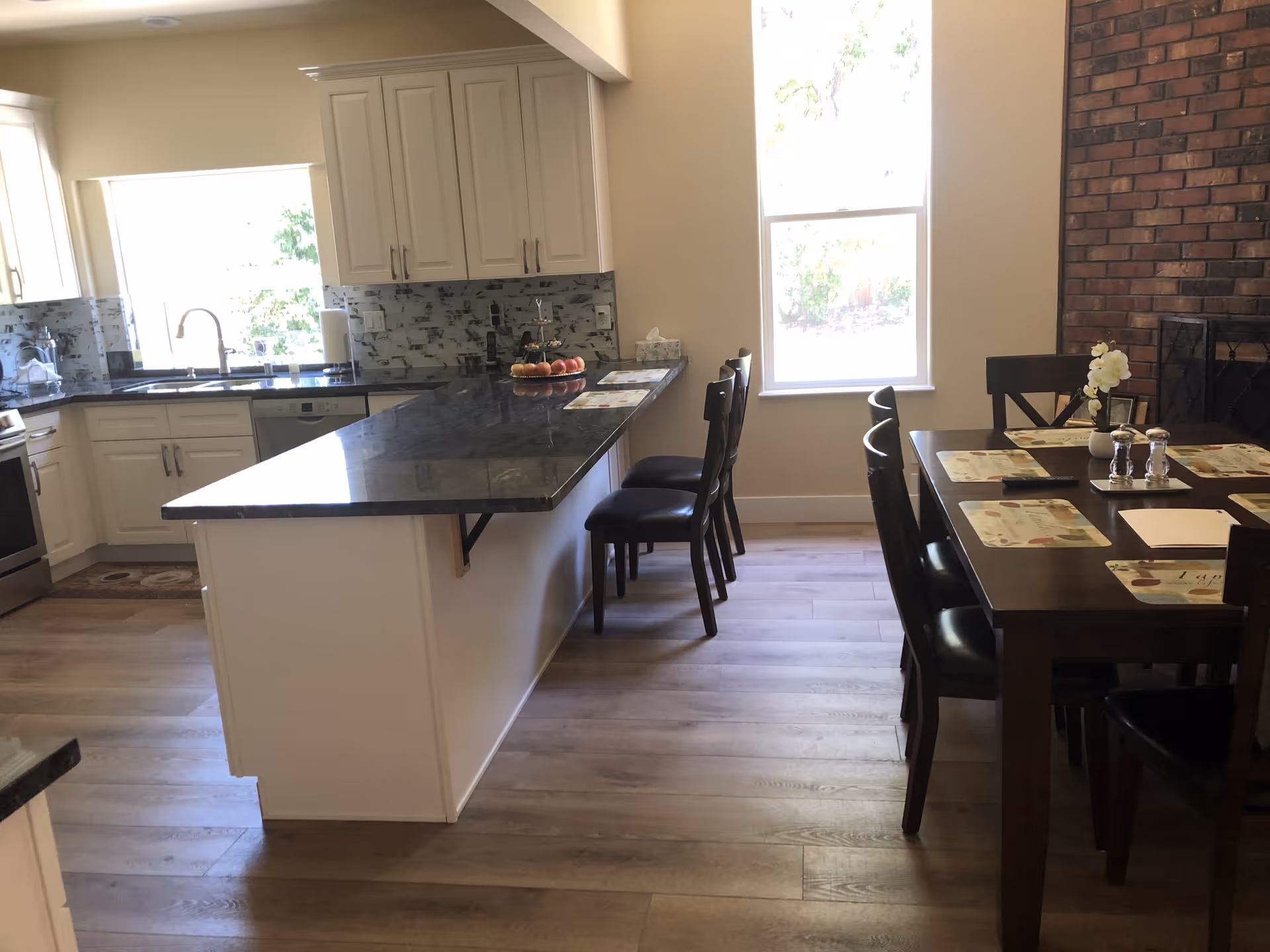 A bright kitchen and dining area with white cabinets, a black granite countertop island with chairs, a dining table set with placemats and chairs, a brick fireplace, and large windows letting in natural light.