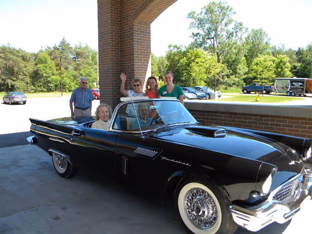 A classic black convertible car parked under a brick archway with four elderly people sitting inside and three adults standing behind the car. The background shows a parking lot with several cars and green trees.