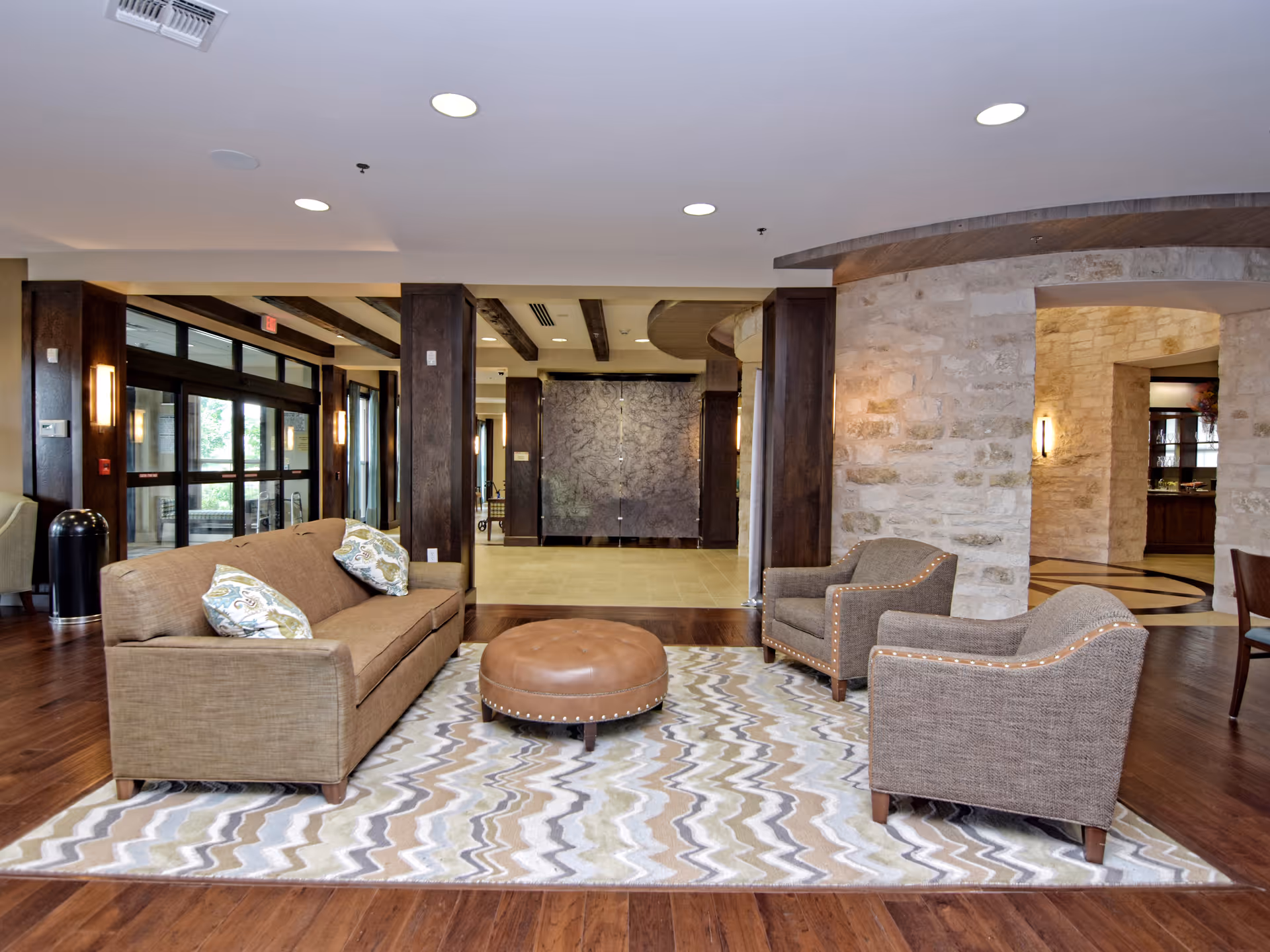 A cozy seating area in a senior living facility lobby featuring a brown sofa with patterned cushions, two matching armchairs, and a round leather ottoman on a patterned rug. The space has wooden flooring, stone walls, and large glass doors leading outside.