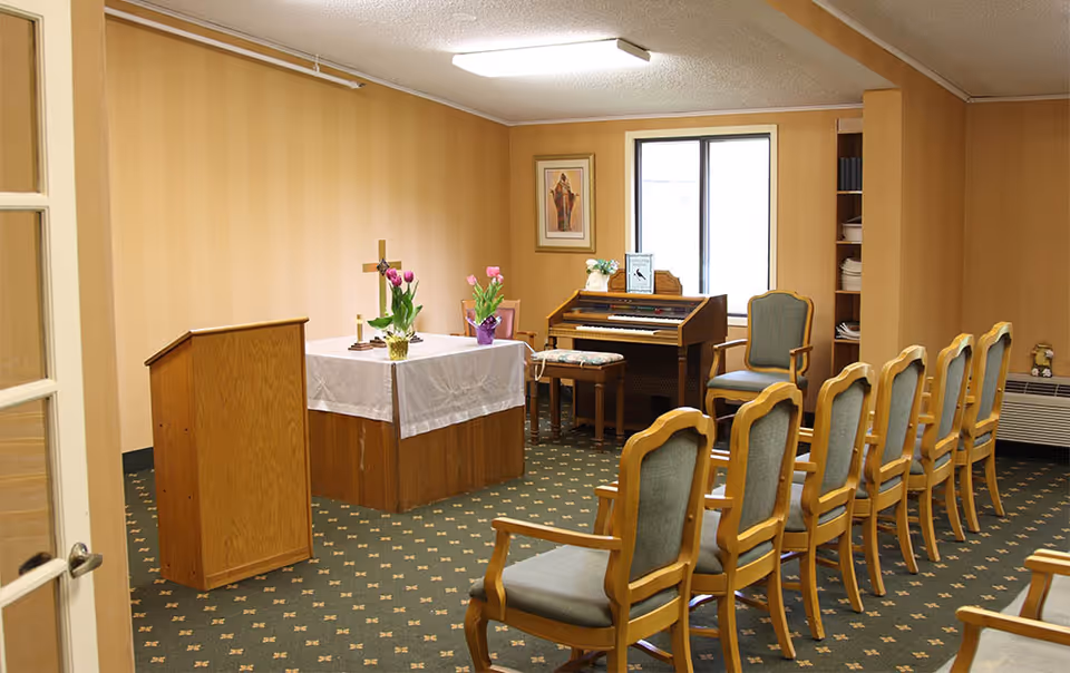 Small chapel room with wooden chairs arranged in rows facing a wooden podium and altar covered with a white cloth and decorated with flowers. There is a cross on the altar, a framed religious picture on the wall, and an organ with a bench near a window.