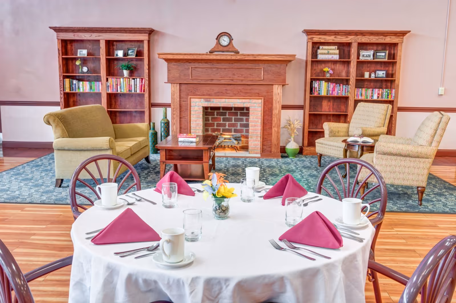 Round dining table set with cups and folded magenta napkins in a communal room with a fireplace, bookshelves, and armchairs.