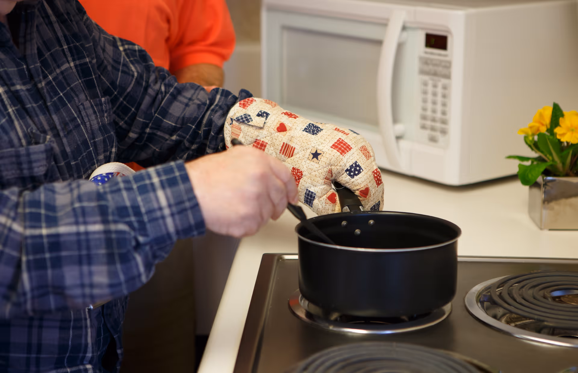 A person wearing a blue plaid shirt and an oven mitt with a patriotic pattern is stirring a black pot on an electric stove. In the background, there is a white microwave and a small plant with yellow flowers on the kitchen counter.