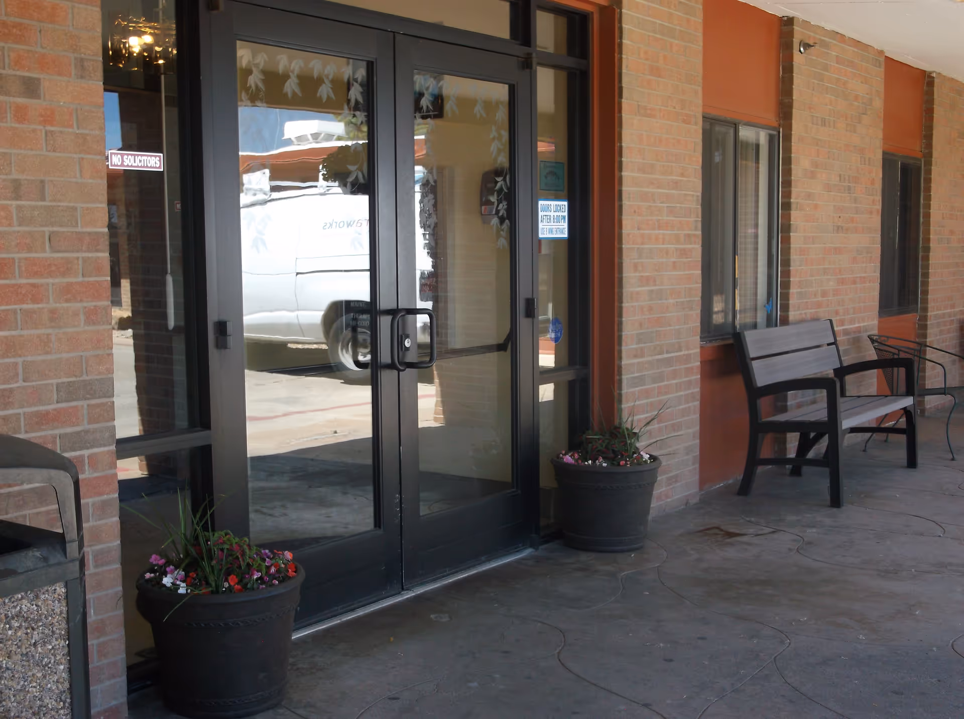 Glass double-door entrance to a brick building with benches and potted flowers on a covered walkway.