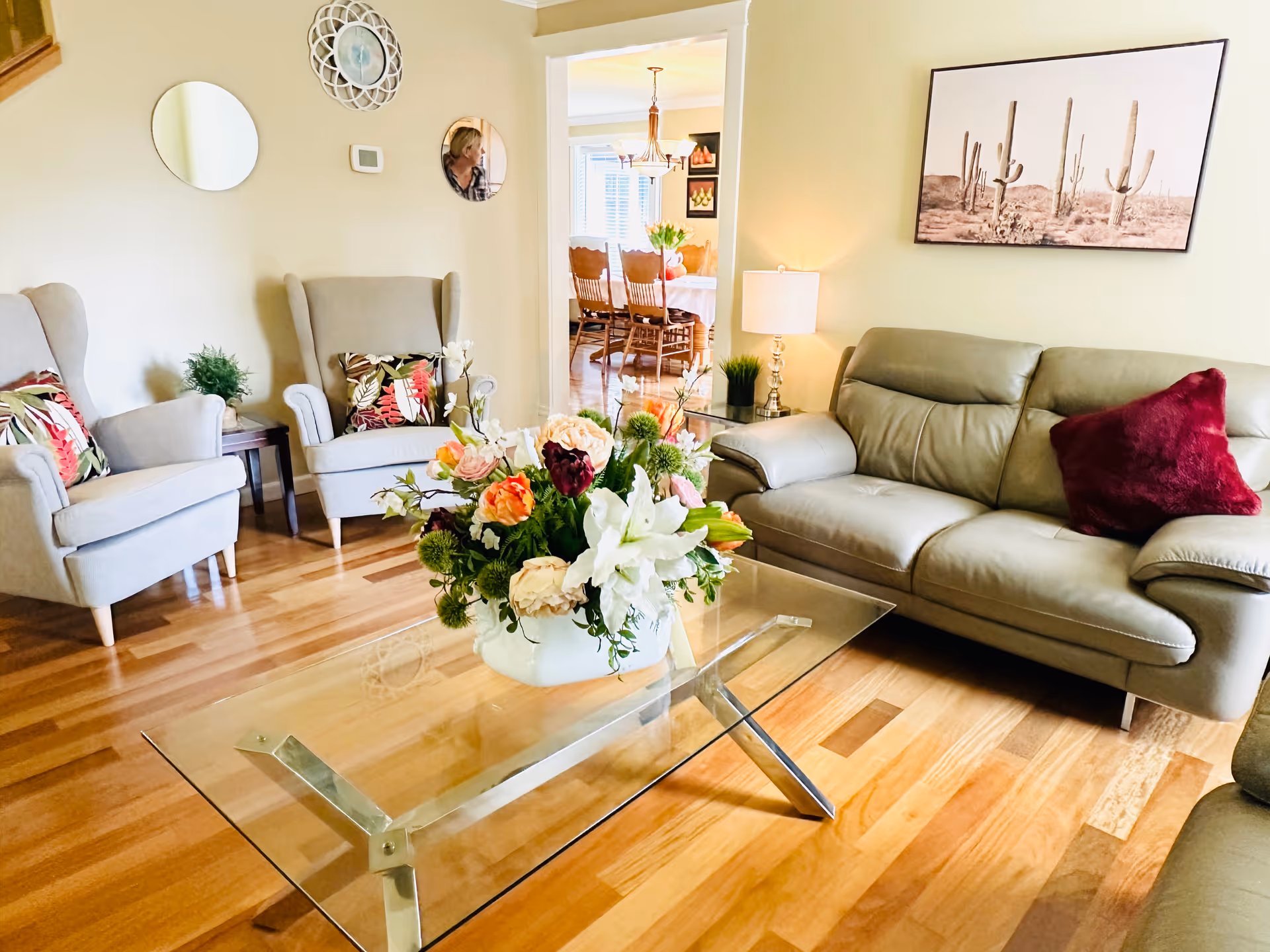 A cozy living room with a gray leather sofa adorned with a red pillow, two gray armchairs with floral cushions, and a glass coffee table with a large floral arrangement. The room has wooden flooring, a side table with a lamp and plant, and a wall decorated with round mirrors and a framed picture of cacti. In the background, a dining area with a wooden table and chairs is visible.