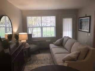 A cozy living room with a beige sofa, a round dark coffee table, a side chair, and a wooden cabinet with a lamp and decorative items. There is a large window with white blinds and a door with a window panel. The walls are light-colored with framed artwork.