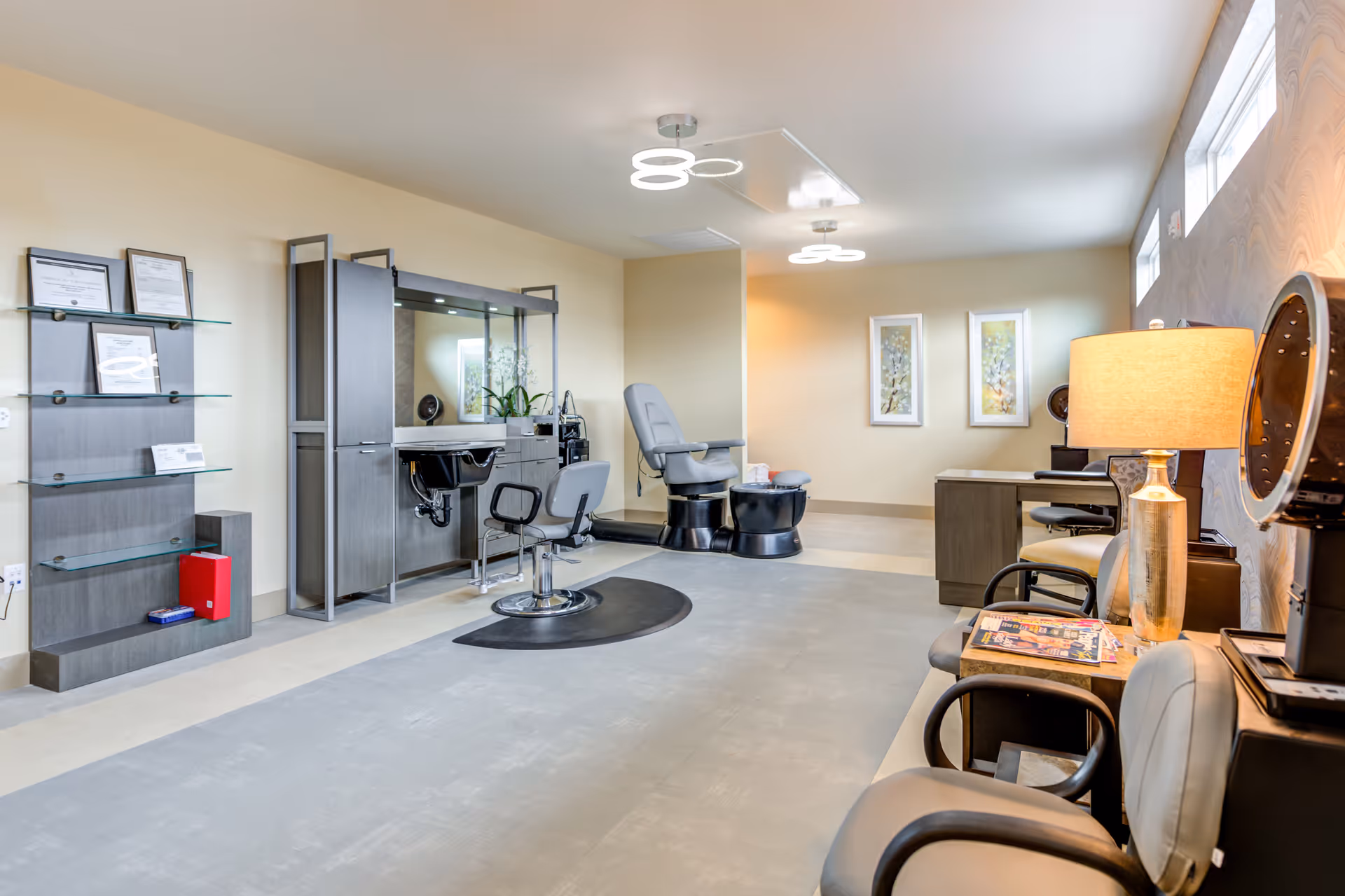 A bright and modern salon area in a senior living facility with styling chairs, a hair washing station, and a pedicure chair. The room has light-colored walls, contemporary lighting fixtures, framed artwork on the walls, and a small waiting area with chairs and a lamp.