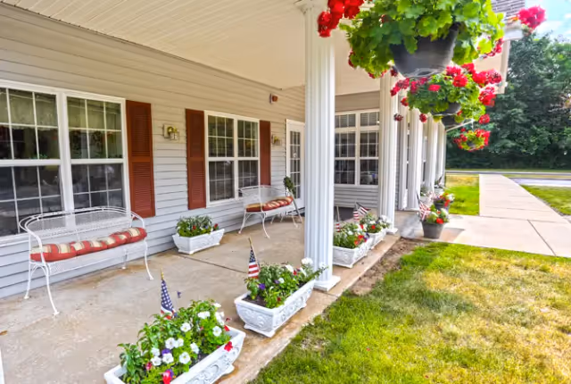 Covered front porch with white columns, benches, hanging flower baskets and planter boxes beside a sidewalk and lawn.