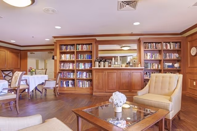 A cozy senior living common area with wooden bookshelves filled with books, a coffee station with cups and dispensers, a glass-top wooden coffee table with a white flower arrangement, beige upholstered armchairs, and round tables with chairs covered in white tablecloths. The room has wooden flooring and soft lighting.