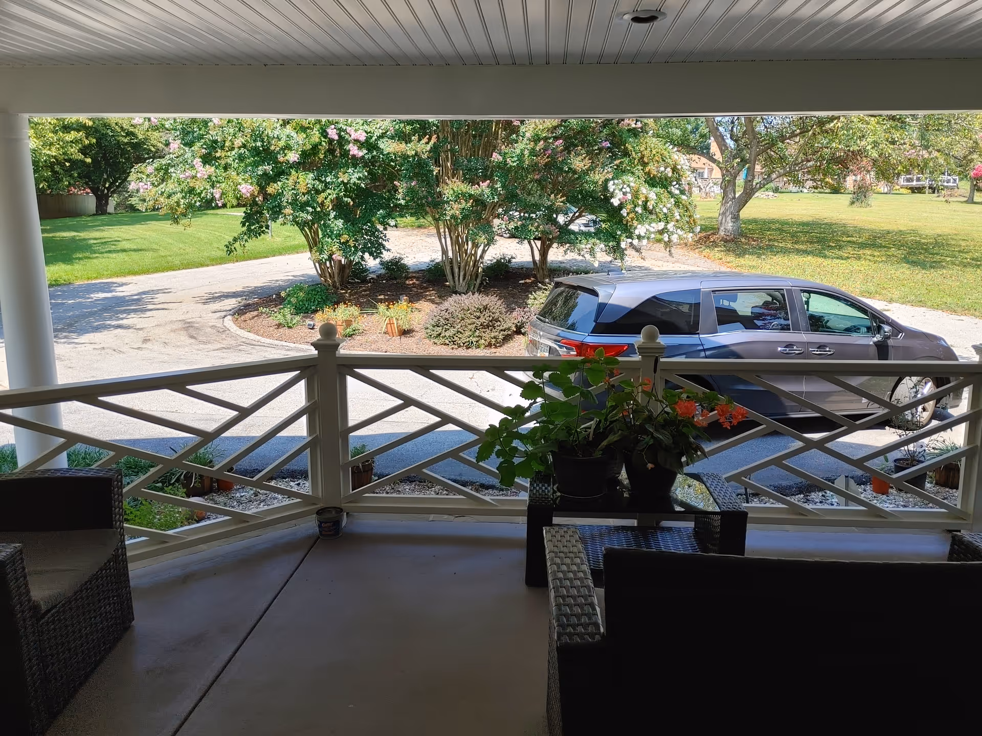 View from a covered porch with wicker chairs and a small table holding potted plants. Beyond the porch railing, there is a driveway with a parked gray minivan and a landscaped area with flowering bushes and trees. A grassy lawn and more trees are visible in the background.