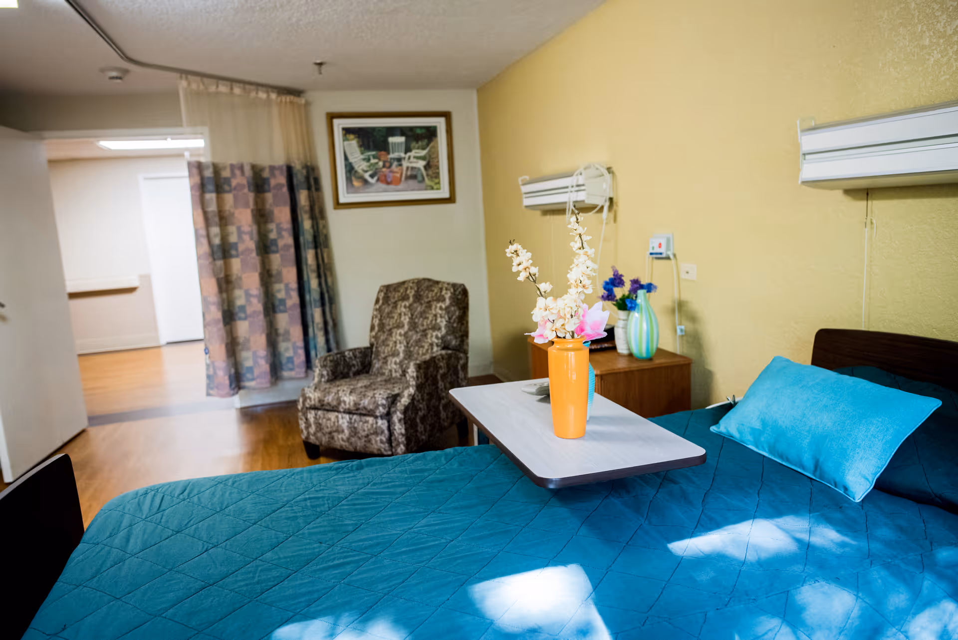 A senior living facility bedroom with a teal bedspread and matching pillow, a small table with a vase of flowers on the bed, a patterned armchair, a wooden nightstand with decorative vases, a framed picture on the wall, and a curtain partially covering a doorway.