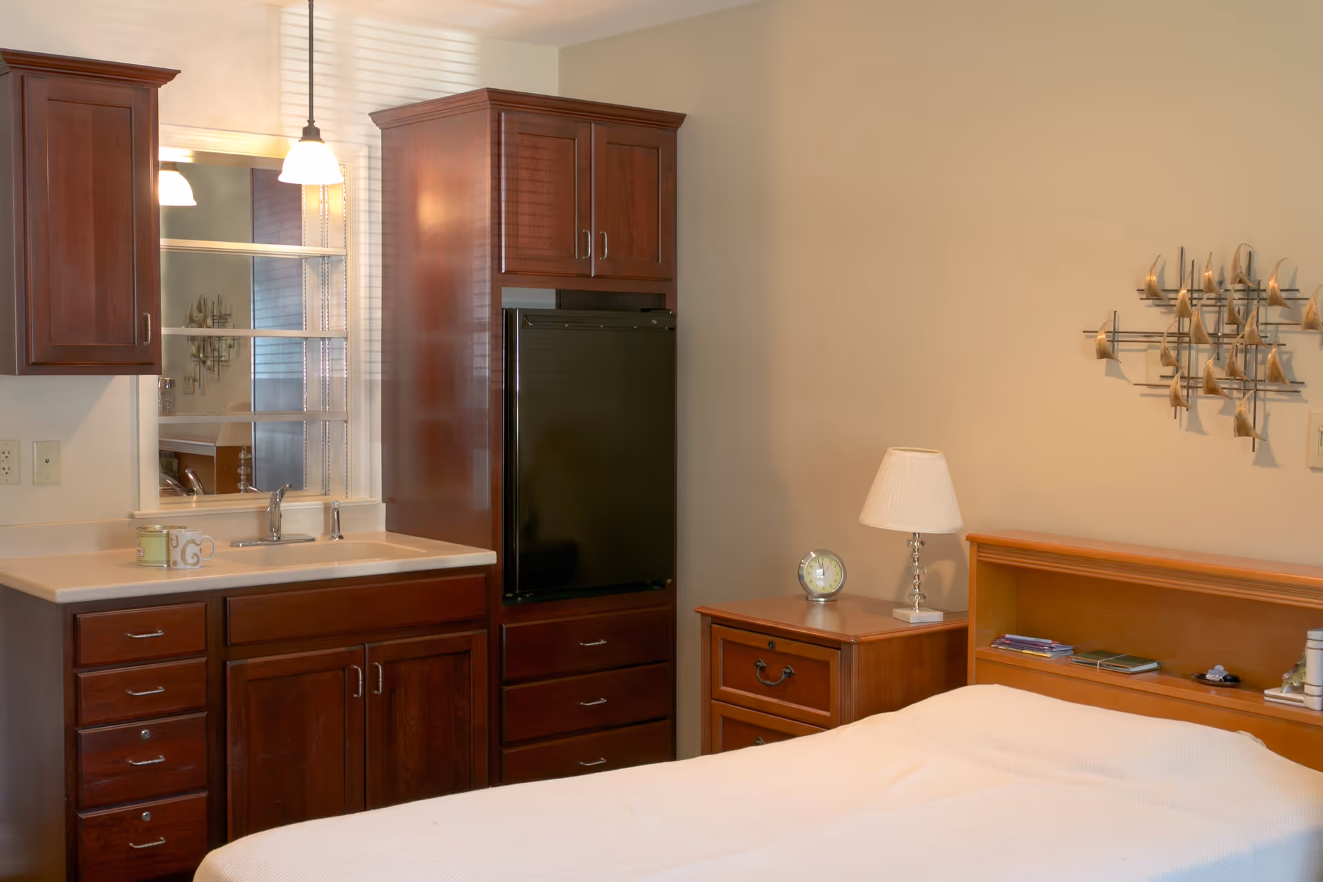 A bedroom with a bed covered in a white blanket, a wooden nightstand with a lamp and clock, and a wooden cabinet with a small refrigerator and a sink with a mirror above it. The walls are beige and there is a decorative metal wall art piece above the bed.
