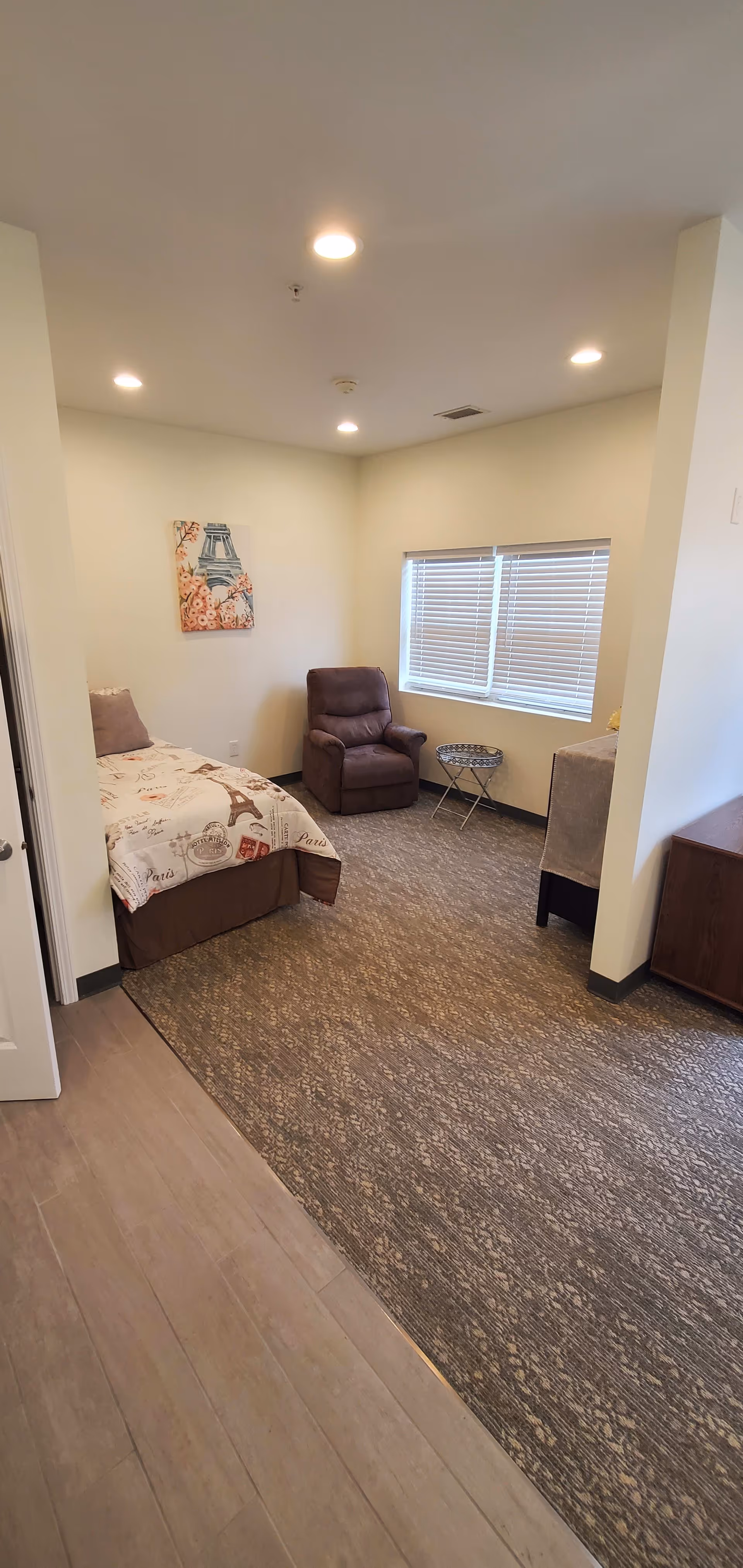 A small bedroom with a single bed covered in Paris-themed bedding, a brown recliner chair, a small round side table, a window with blinds, and a piece of wall art featuring the Eiffel Tower and flowers. The room has beige walls, recessed ceiling lights, and a carpeted floor with a wood-like floor section near the entrance.