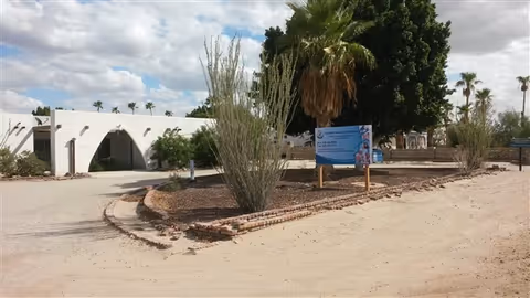 Single-story white stucco building with arched entrance, desert landscaping, palm trees, and a freestanding sign for Foothills Adult Care Home.