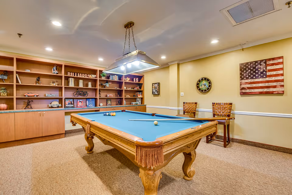 A well-lit room with a billiards table in the center, featuring a blue felt surface and billiard balls arranged on it. Behind the table is a large wooden shelving unit with various decorative items, including a model bicycle, books, and a sign that reads 'BILLIARDS'. Two wooden chairs with woven backs are placed against a beige wall, which is decorated with a clock and an American flag artwork.