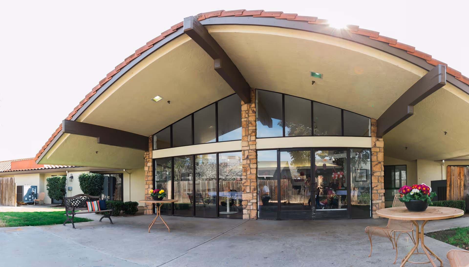 Exterior view of Riverwood Healthcare Center featuring a covered patio area with a curved roof supported by stone pillars. The patio has glass doors and windows, two round tables with flower pots, and a bench with colorful cushions. The sun is shining brightly in the background.