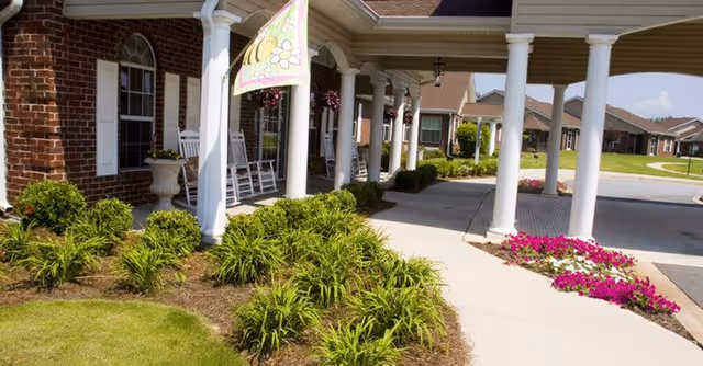 Covered entrance area of a senior living facility with white columns, rocking chairs on the porch, a decorative flag with a bee and flower, well-maintained green shrubs, and pink flowers along the sidewalk.