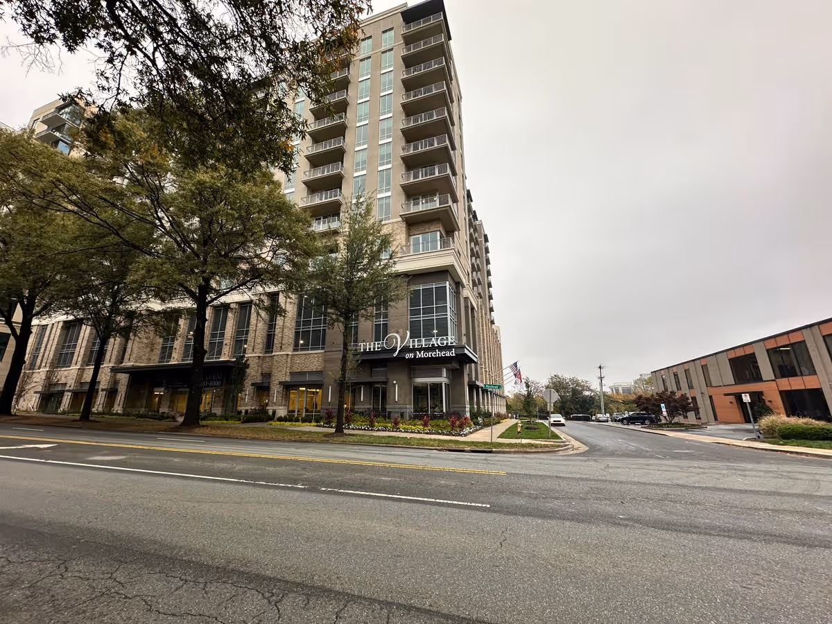 Street view of the multi-story "The Village on Morehead" building with trees, sidewalk, and a road in front.