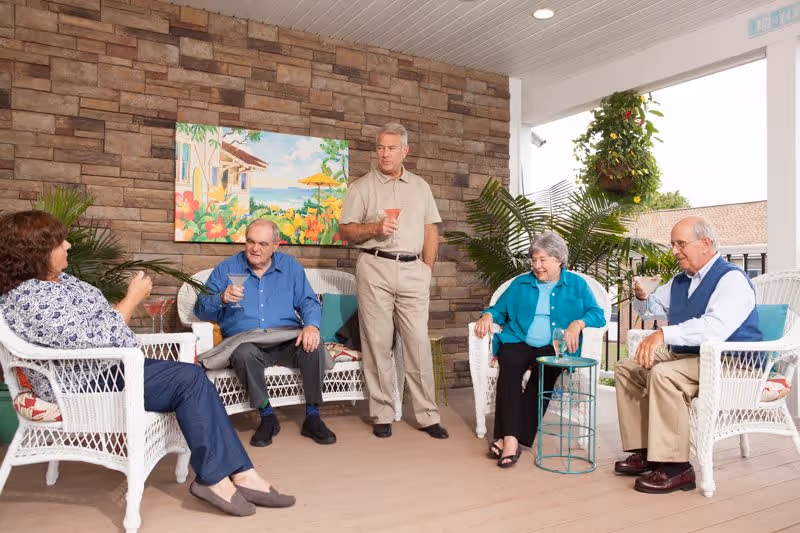 Four elderly people sitting and standing on a covered patio with wicker furniture, holding drinks and engaging in conversation. The patio has a stone wall with a colorful painting, potted plants, and an open view to the outside.