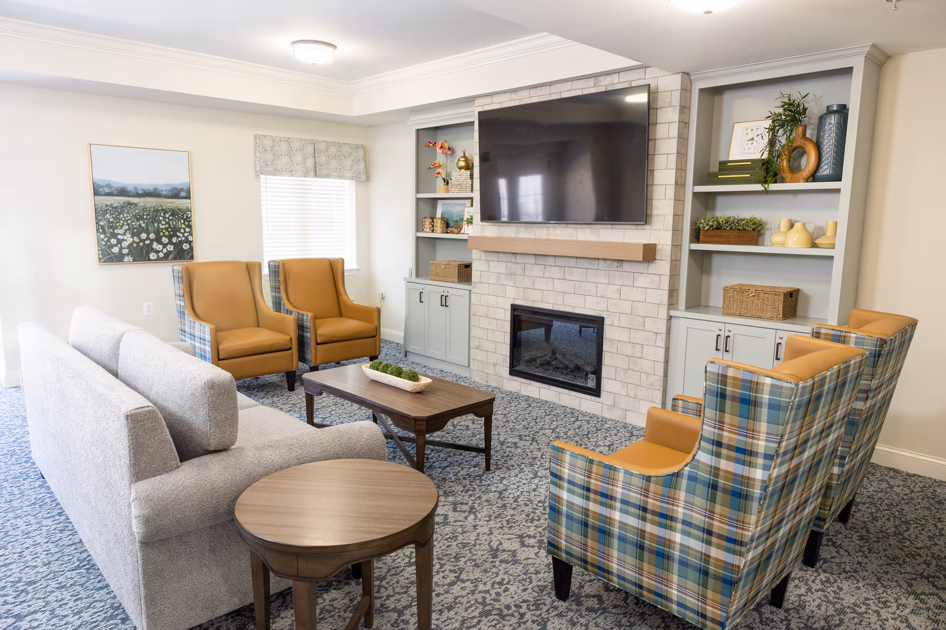 A cozy senior living common area with a gray sofa, two mustard yellow armchairs, and three plaid armchairs arranged around a wooden coffee table. The room features a large flat-screen TV mounted above a stone fireplace with built-in shelves on either side, decorated with plants, books, and vases. A window with a patterned valance and a framed landscape painting are visible on the wall.