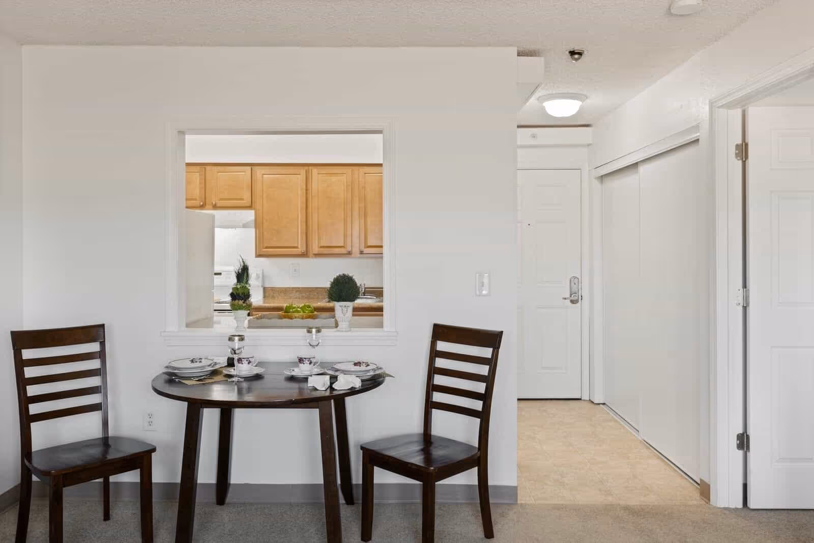 Small dining area with a round dark wooden table set for two with plates, cups, and napkins. Two matching wooden chairs are placed on either side of the table. A pass-through window reveals a kitchen with light wood cabinets, a white refrigerator, and a countertop with decorative plants and green apples. To the right, a hallway leads to a white door and a closet with sliding doors.