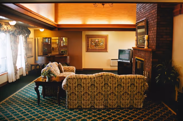 A cozy living room with patterned sofas, a wooden coffee table with a flower vase, a brick fireplace with a mirror and candle on the mantel, a small TV on a stand, framed artwork on the wall, and large windows with floral curtains letting in natural light.