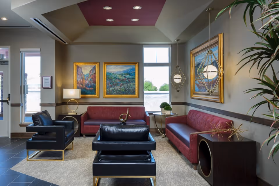A stylish and comfortable living room area with two red sofas, two black leather chairs with gold accents, a beige rug, and modern geometric pendant lights. The walls are decorated with framed landscape paintings, and there are side tables with decorative items and a lamp. Large windows allow natural light to brighten the space.