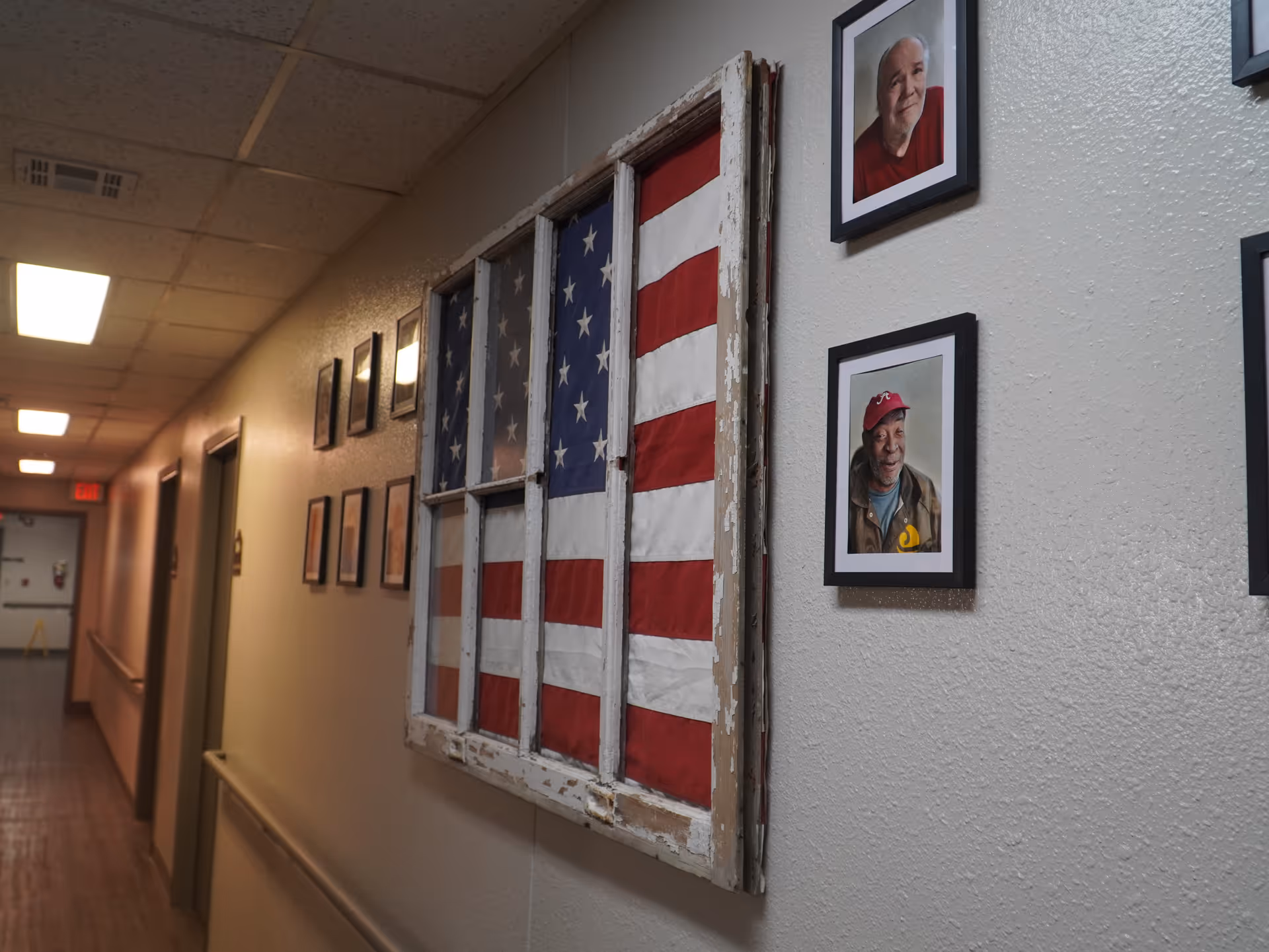 A hallway in an assisted living facility with framed photographs of residents on the wall and a rustic window frame displaying an American flag. The hallway has a handrail and several doors along the left side.