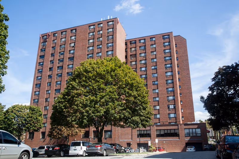 Brick multi-story nursing center building with large trees and parked cars in front under a blue sky.