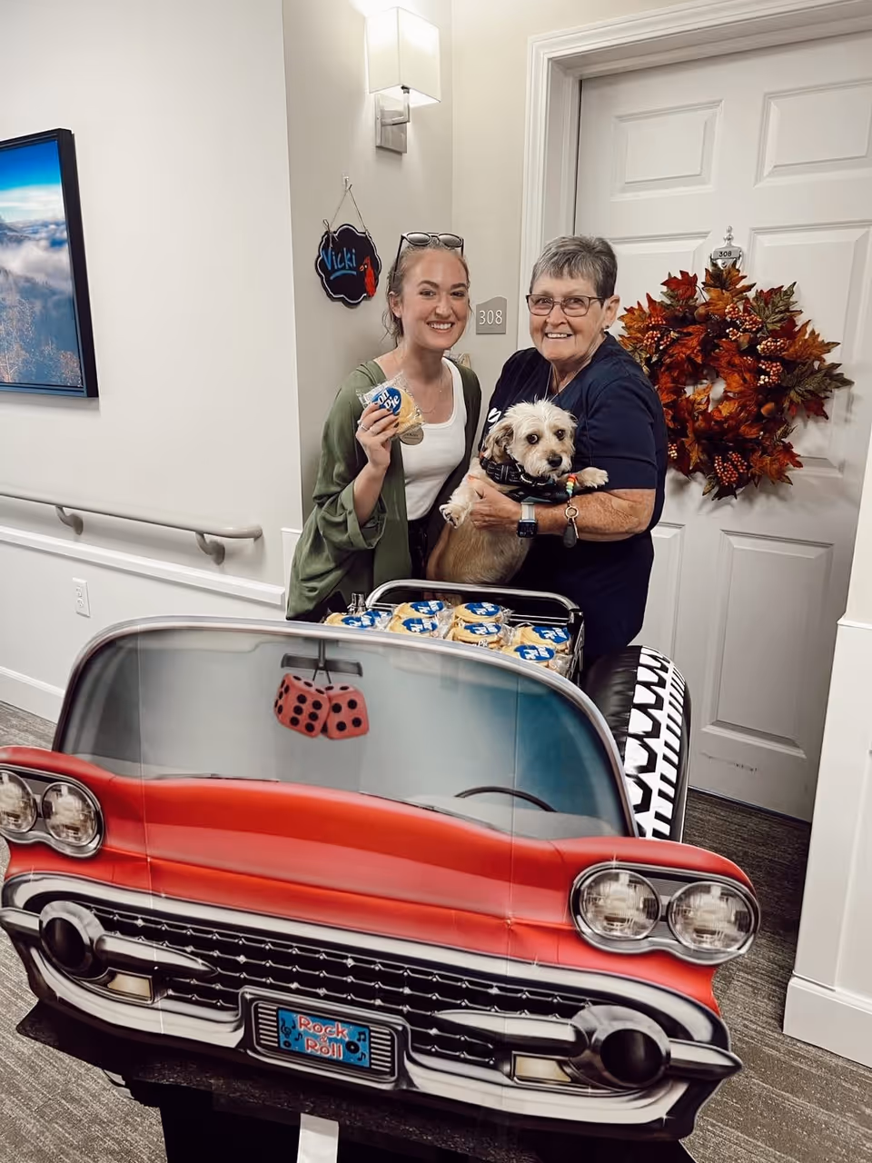 Two women standing in a hallway of a senior living facility, one holding a small dog and the other holding a packaged cookie. They are behind a decorative cart designed to look like the front of a red vintage car with a 'Rock & Roll' license plate and fuzzy dice hanging from the rearview mirror. A door with the number 308 and a fall-themed wreath is visible in the background.