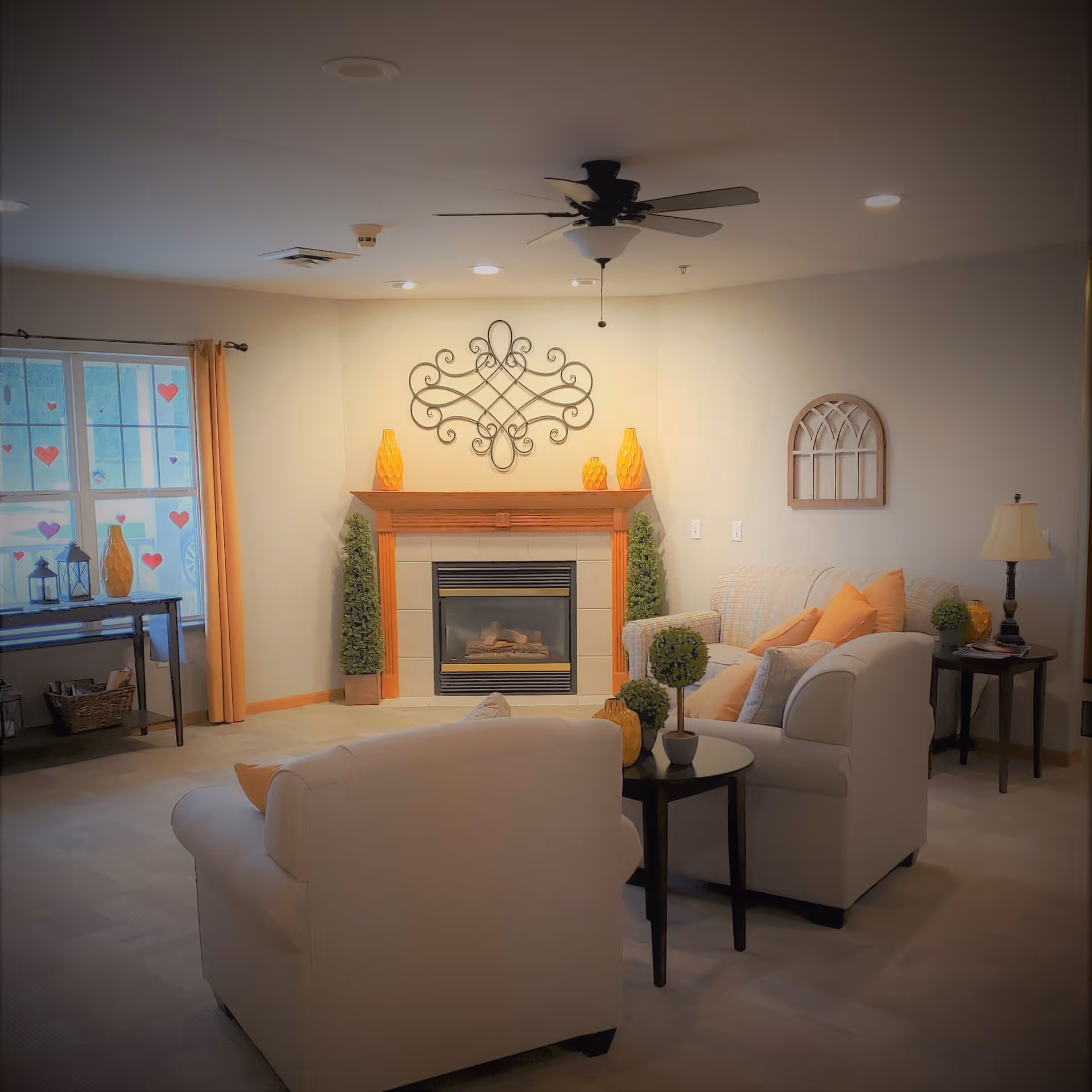 A cozy living room with a fireplace centered on the far wall, decorated with two tall green plants and three orange vases on the mantel. Two cream-colored armchairs and a matching sofa with orange and beige cushions are arranged around a small black coffee table with decorative plants. A ceiling fan with lights hangs above, and a window with orange curtains and heart-shaped decorations is on the left side. A side table with a lamp and more decor is on the right.