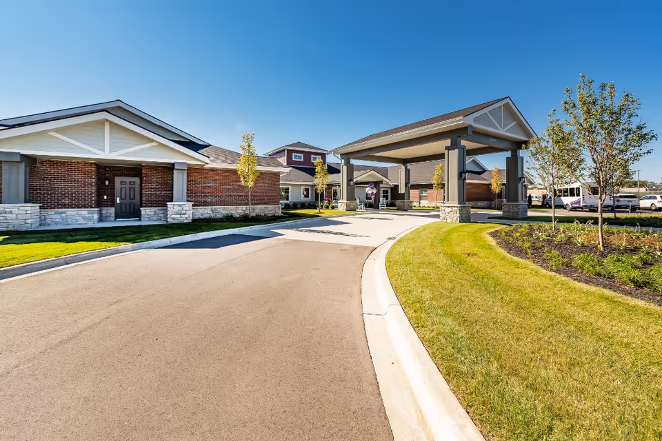 Exterior view of Provision Living at Livonia showing a driveway leading to a covered entrance with brick and stone buildings on either side, surrounded by green lawns and small trees under a clear blue sky.