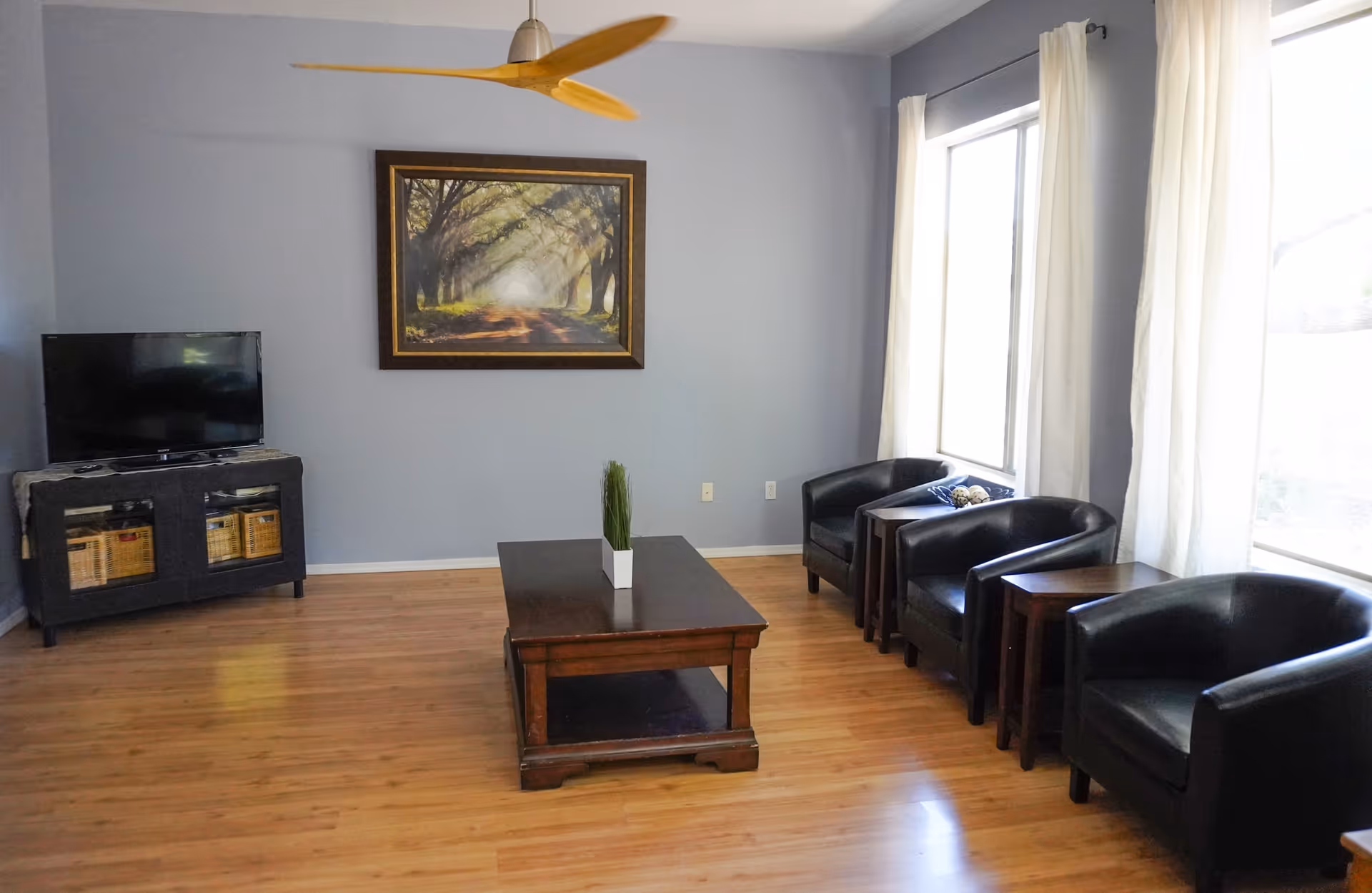 Bright living room with hardwood floors, a central wooden coffee table, a TV on a stand, framed artwork on the wall, and a row of black armchairs by large windows.