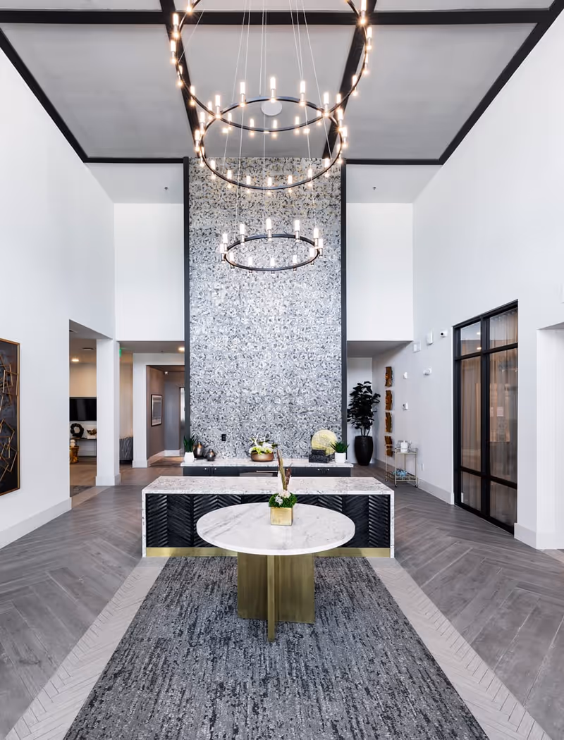 A modern and elegant interior space with high ceilings featuring two large circular chandeliers. In the center, there is a round marble table with a small plant on top, placed on a textured gray rug. Behind the table is a long rectangular marble counter with decorative plants and objects. The back wall has a decorative patterned design, and the floor is covered with light gray wood-like tiles arranged in a herringbone pattern. The space has white walls and black trim accents, with doorways and windows visible on the sides.