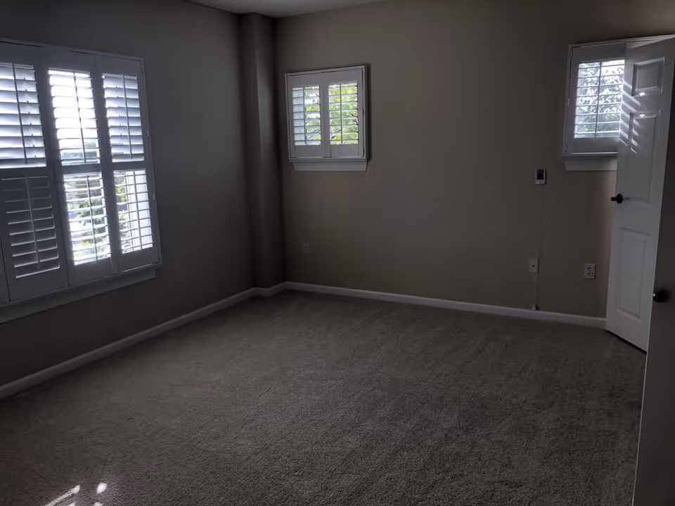 Empty room with beige carpet, beige walls, three windows with white plantation shutters, and a white door partially open on the right side.