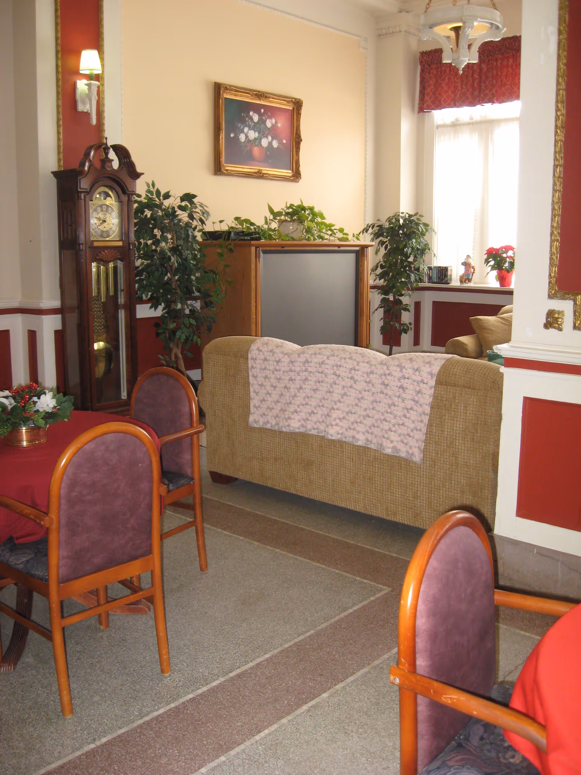 A cozy living room area with a beige couch draped with a pink patterned blanket, two purple cushioned wooden chairs around a table with a red tablecloth, a grandfather clock, potted plants, a painting of flowers on the wall, and a window with red valance curtains letting in natural light.
